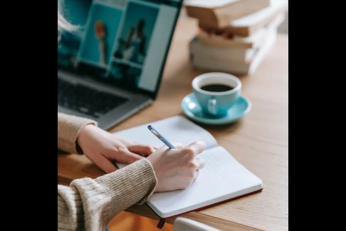 Someone writing in a notebook beside a laptop and a cup of coffee